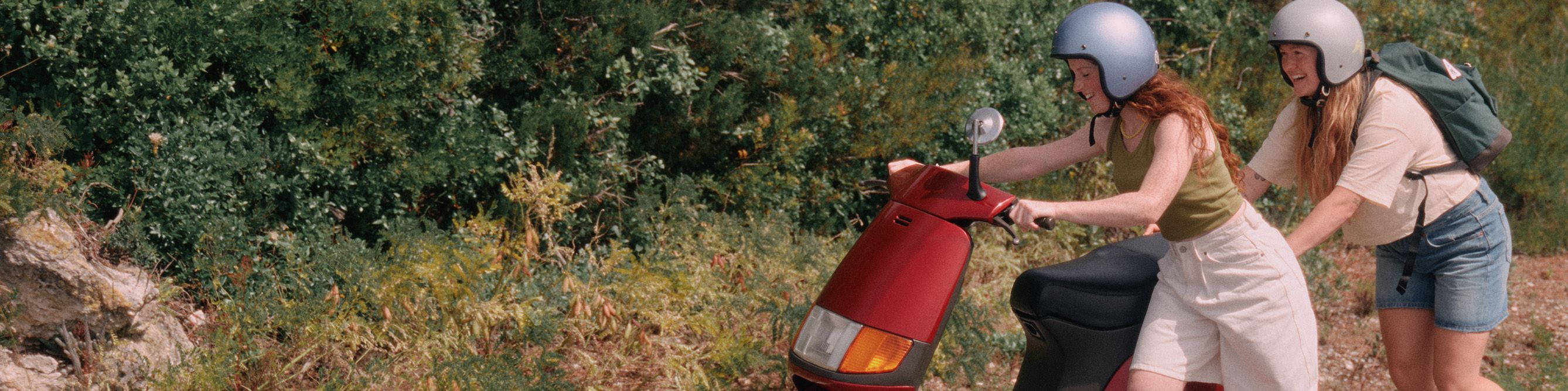 Zwei Freundinnen schieben einen roten Motorroller eine Landstraße entlang. 