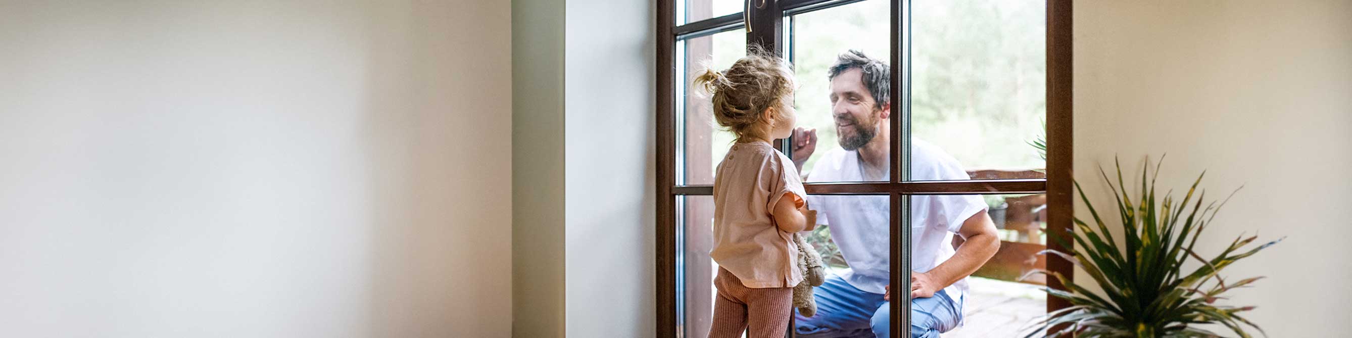 Vater und kleine Tochter schauen sich durch Fensterscheibe an und lächeln