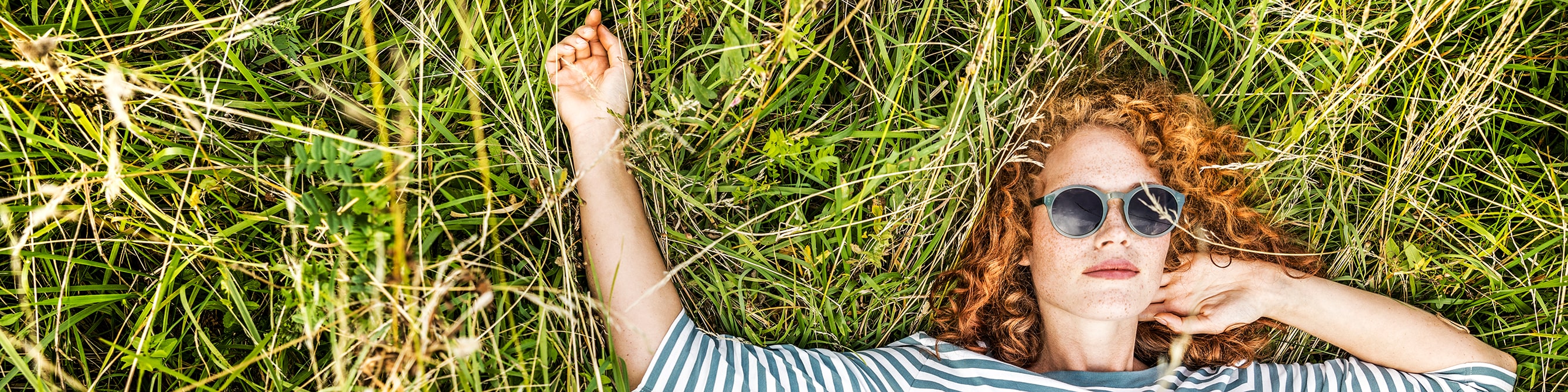 Frau mit Sonnenbrille und roten Locken liegt auf dem Rücken auf einer Wiese.