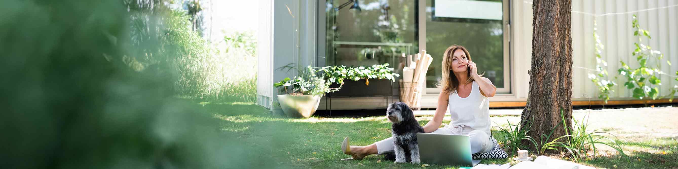 Frau sitzt mit Hund, Laptop und Handy auf einem Kissen im Garten vor ihrem Haus
