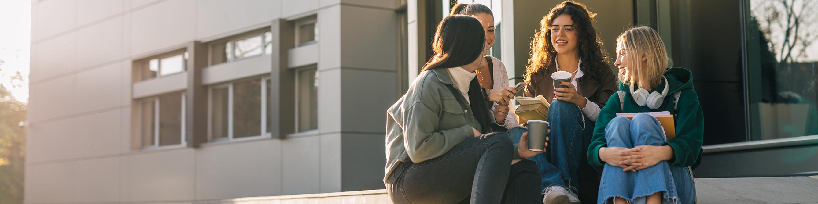 Vier junge Frauen sitzen auf einer Treppe, trinken Kaffee aus Pappbechern und unterhalten sich angeregt.