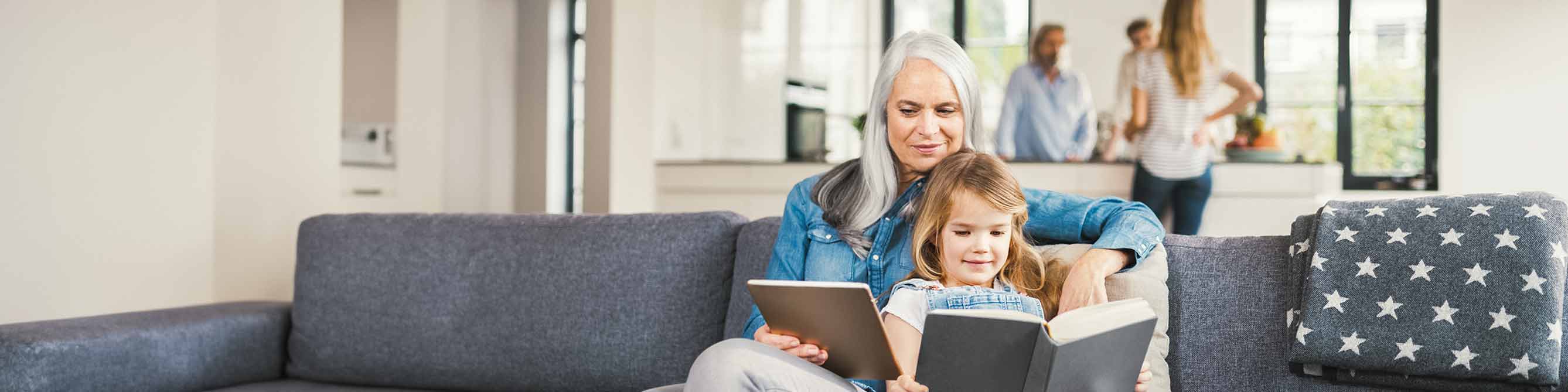 Eine Großmutter und ihre Enkelin sitzen auf einem Sofa, beide lesen parallel auf einem Tablet PC und in einem Buch.