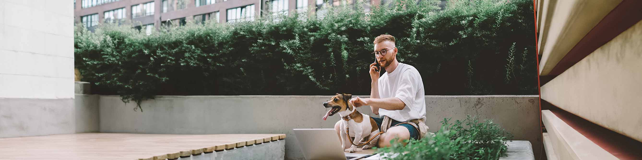 Mann sitzt draußen und telefoniert, mit einer Hand hält er seinen Hund am Halsband fest 
