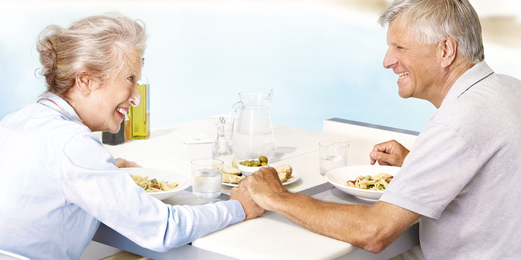 Ein älteres Ehepaar sitzt am Frühstückstisch, es hält sich lächeln die Hand.