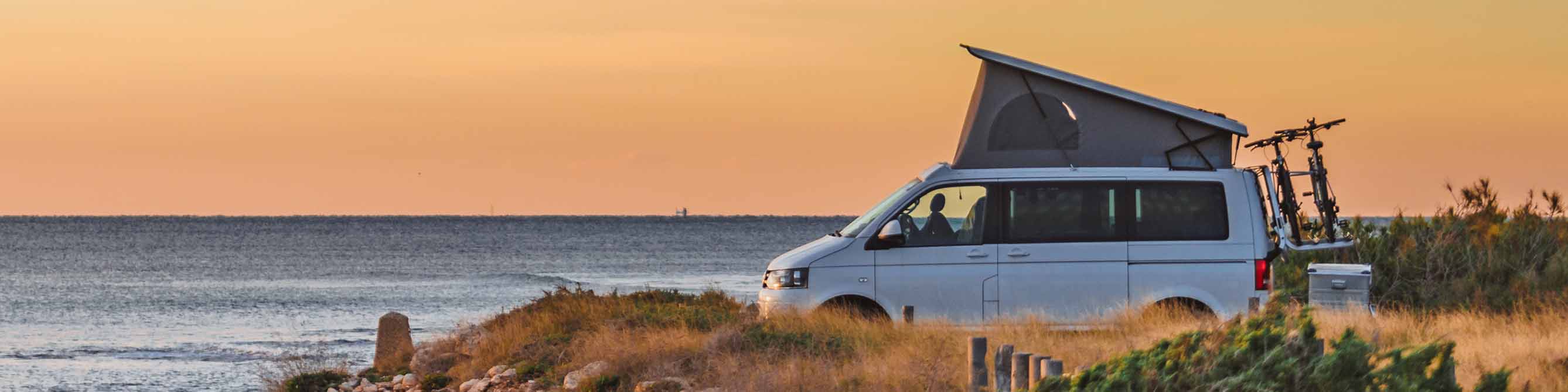Ein weißer Camper-Van steht an einer Küste mit Blick auf das Meer und den Sonnenuntergang. 