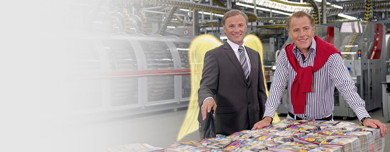 Ein Schutzengel im Anzug und ein Druckerei-Mitarbeiter stehen vor einem Stapel mit Druckerzeugnissen.