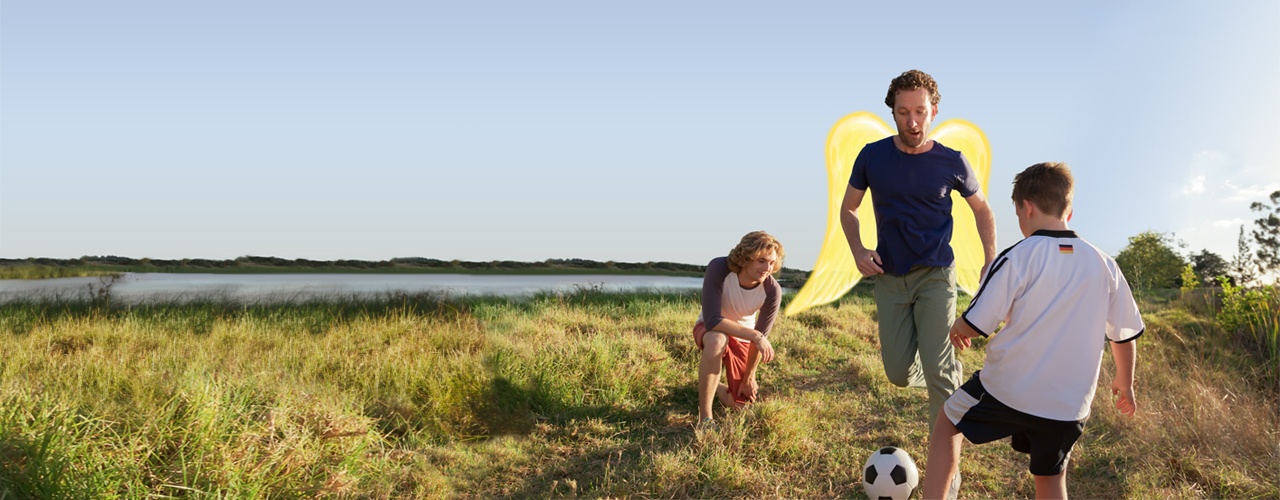 Private Unfallversicherung: Ein Vater spiel mit seinen Kindern Fußball. Der Vater hat Schutzengel-Flügel.